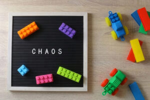 Flat lay of a black letter board with the word “CHAOS” and scattered colorful LEGO-style bricks on a wooden table, with toy cars and geometric blocks on the right.